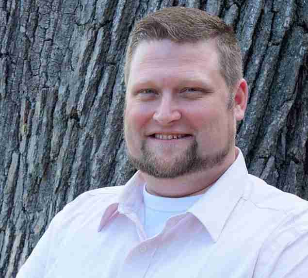 a man with short, neatly trimmed hair and a beard. He is wearing a light pink button-up shirt over a white t-shirt. The man is smiling and standing in front of a tree with a textured bark, which forms the background of the image. The setting appears to be outdoors, suggesting a natural and relaxed environment.